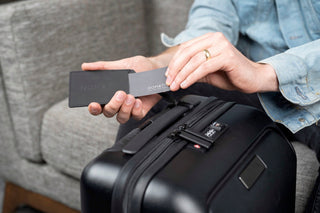 NOMATIC Accessory Best Sellers collection banner, close-up of hands attaching a branded luggage tag to a black suitcase