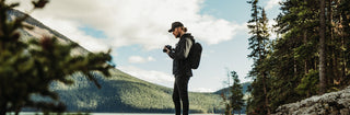 NOMATIC All Camera collection banner, man standing near a lake holding a camera, wearing a camera backpack