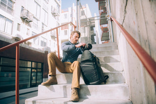 NOMATIC Camera Bags Best Sellers collection banner, man sitting on outdoor stairs beside a black camera backpack, holding a camera