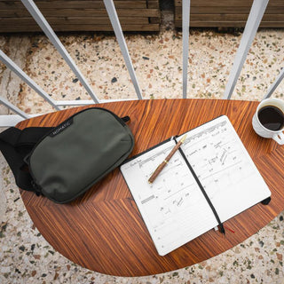 NOMATIC Everyday Accessories collection banner, olive sling bag resting on a wooden table beside an open notebook and a cup of coffee
