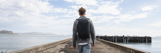 NOMATIC Backpacks collection banner, man wearing a gray backpack walking along a pier over water