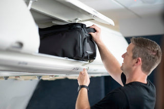 NOMATIC Travel Bags collection banner, man stowing a black travel bag in an airplane overhead bin