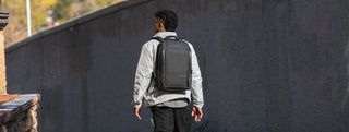 NOMATIC All Backpacks & Bags collection banner, man walking along a concrete wall wearing a gray backpack
