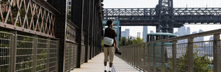 NOMATIC Sling Camera Bags collection banner, man walking across a bridge walkway wearing a tan sling bag and carrying a camera