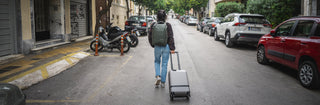 NOMATIC Carry-On Travel Bags collection banner, person walking down a city street pulling gray carry-on luggage