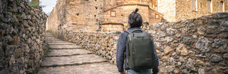 NOMATIC Packs & Bags Best Sellers collection banner, man walking along a stone wall path wearing an olive green backpack
