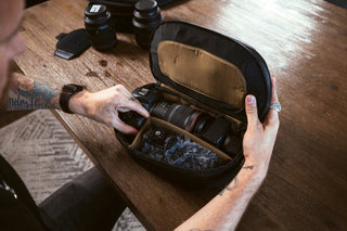 NOMATIC Camera Accessories collection banner, hands packing a camera case organized with lenses and binoculars