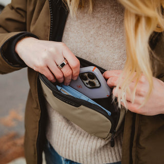 NOMATIC All Accessories collection banner, close-up of a hand unzipping a tan sling bag with a phone tucked inside